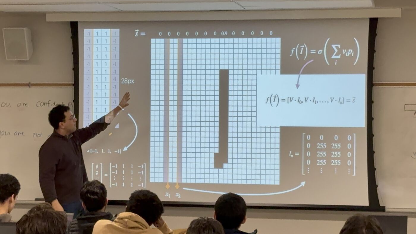Image of person giving lecture in Exam hall with diagrams projected onto a wall.